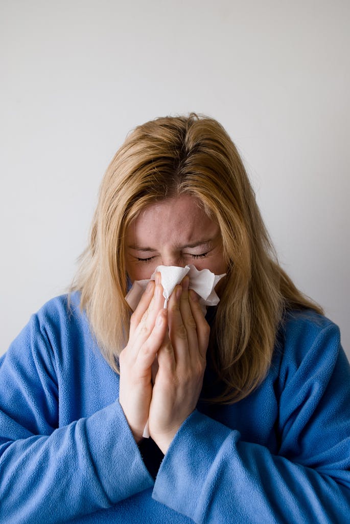 A woman in a blue robe is sneezing into a tissue, due to allergies